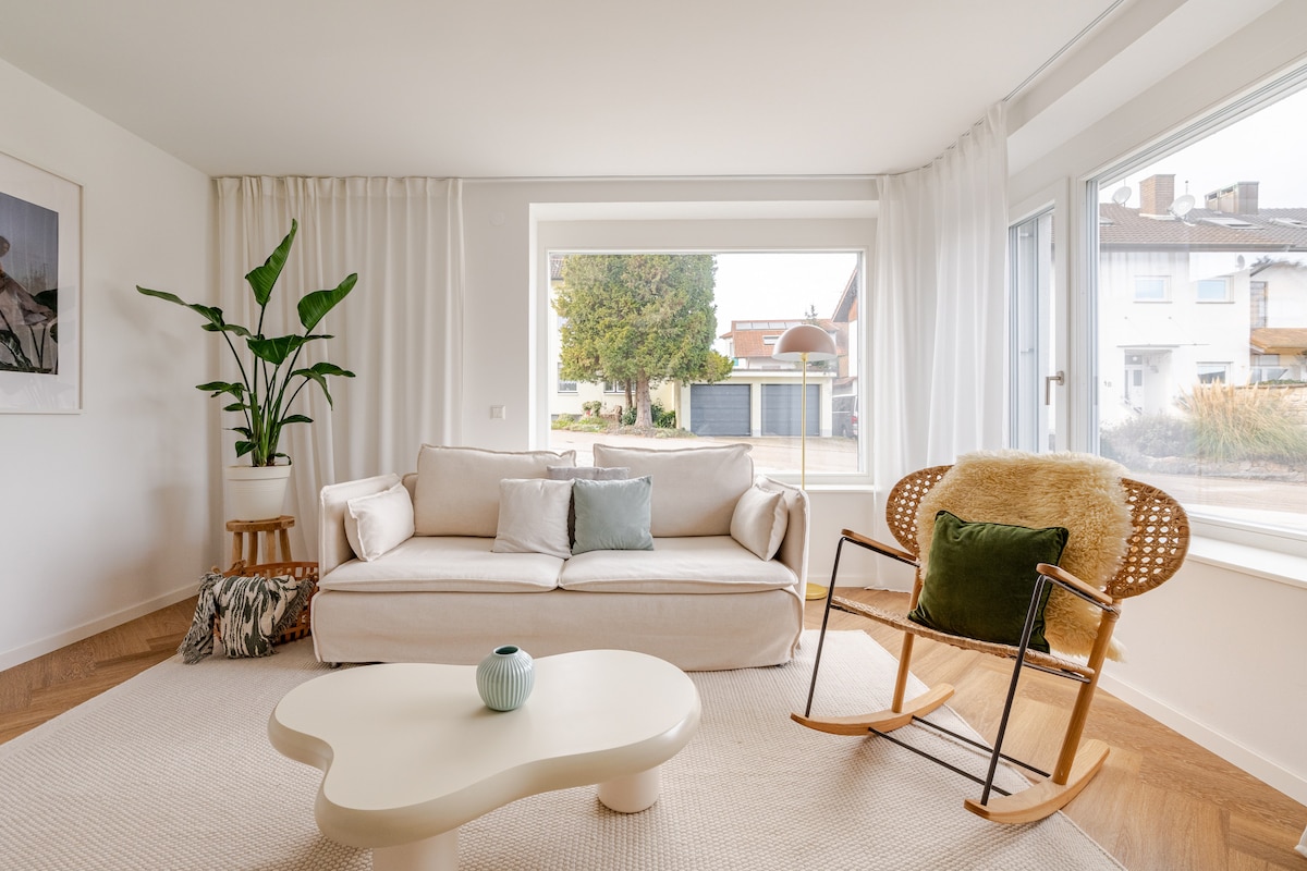 A light-filled living space features a comfortable sofa adorned with decorative cushions. A cozy rocking chair and a unique coffee table are positioned on a textured rug. Greenery from a potted plant adds a natural touch near the window, enhancing the inviting ambiance.