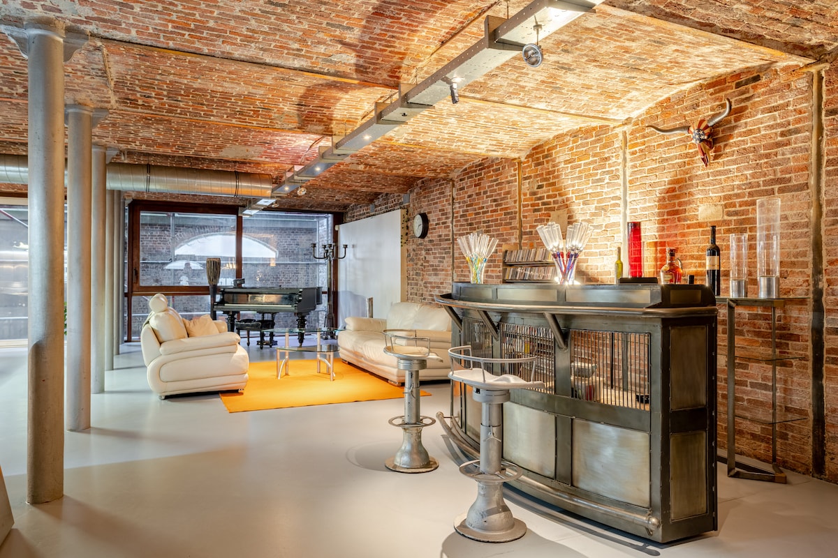 An open area features exposed brick walls, enhancing a rustic ambiance. A bar with stylish stools is complemented by a cozy seating arrangement on a vibrant orange rug. Large windows allow natural light to fill the space, creating an inviting atmosphere.