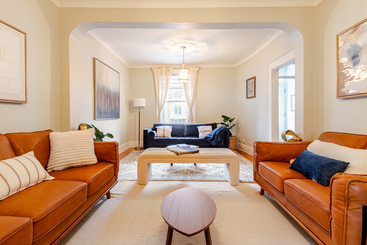 A spacious living room features two stylish leather sofas, a wooden coffee table, and a light area rug. Natural light enters through large windows, illuminating the room's neutral tones and decorative elements. Plants are placed around the space, adding touches of greenery.