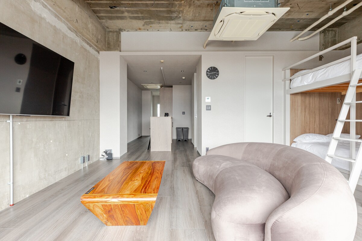 A modern living area features a unique curved sofa in a soft hue, accompanied by a wooden coffee table. A large television is mounted on the wall, while warm natural light fills the space through windows. A minimalist dining area with stools can be seen in the background.