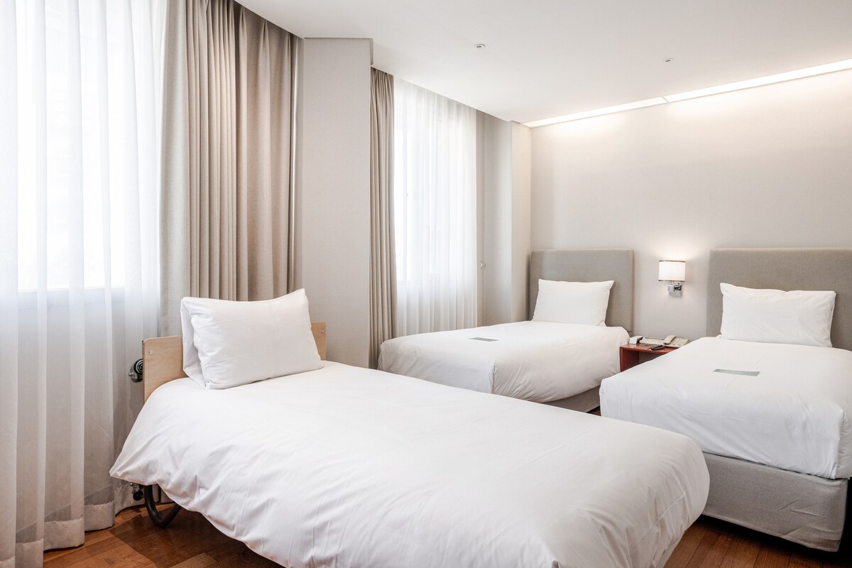 A well-lit guest room features two comfortable beds, each adorned with white linens. Sheer curtains allow natural light to filter through, softly illuminating the neutral-toned walls. A small nightstand is positioned beside one bed, enhancing the room's functionality and welcoming ambiance.