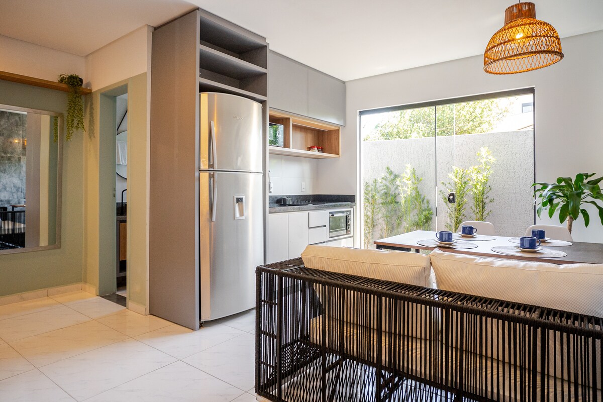 A modern kitchen and dining area features sleek cabinetry and a spacious table set for four. Natural light enters through large windows, illuminating the stylish decor. A refrigerator and microwave are present, with greenery peeking through the glass doors leading to a small outdoor space.