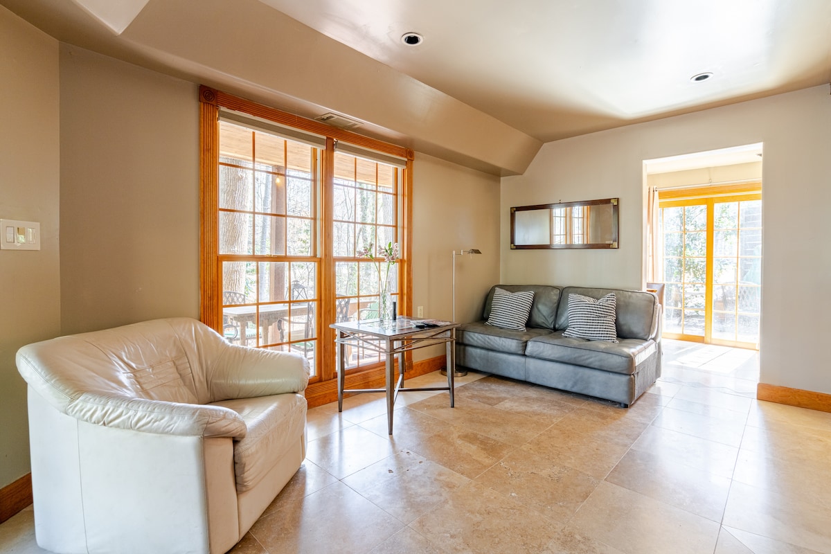 The bright living area features large windows that allow natural light to fill the space. A comfortable sofa and a white armchair provide seating, alongside a glass table. The room's tile flooring contributes to a clean and open feel, leading to a patio view.