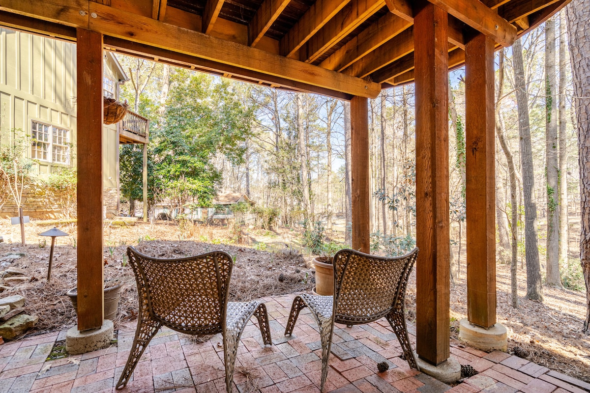 An outdoor patio area is framed by wooden beams, featuring two woven seating chairs placed on a patterned brick surface. The surrounding greenery provides a natural backdrop, creating a serene environment. Nearby elements include potted plants and hints of a wooded landscape.