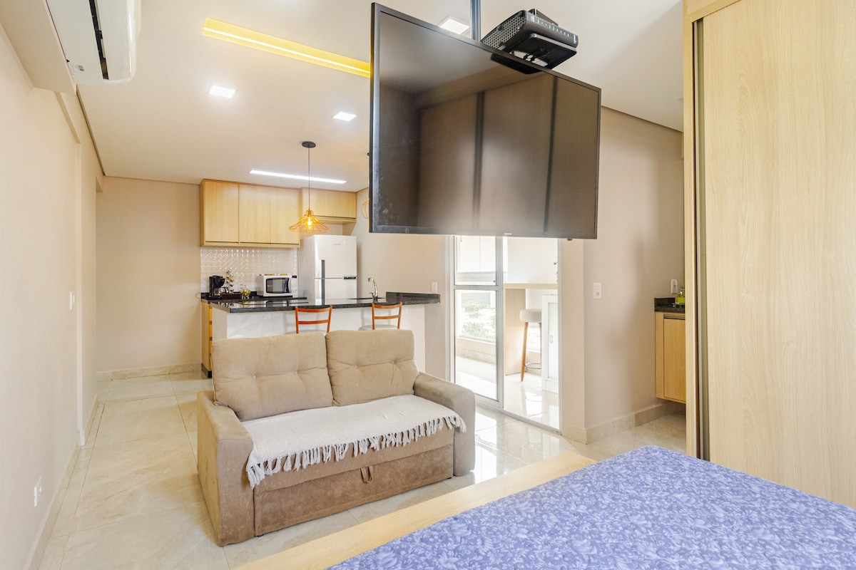 A charming living area features a light beige sofa draped with a white throw. A flat-screen TV is mounted on a wall, positioned in front of a modern kitchen with wooden cabinetry and stainless steel appliances. Bright light filters through a sliding glass door leading outside.