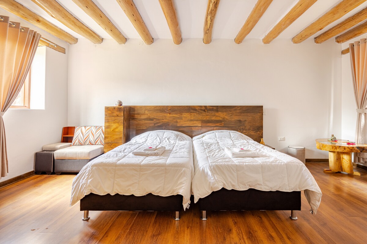 A bright and spacious bedroom is displayed, featuring two bedside beds with quilted white covers. The warm wooden floor contrasts with the light walls, while natural light enters through a window adorned with beige curtains. A small seating area is visible to the side.