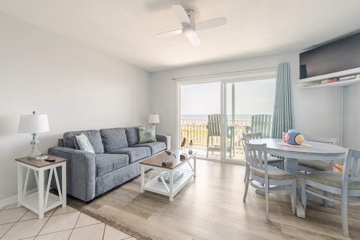 A light-filled living area features a comfortable gray sofa facing a sliding glass door that opens to an oceanfront balcony. A round dining table with light-colored chairs is situated nearby. A ceiling fan and TV are present, enhancing the overall coastal feel.