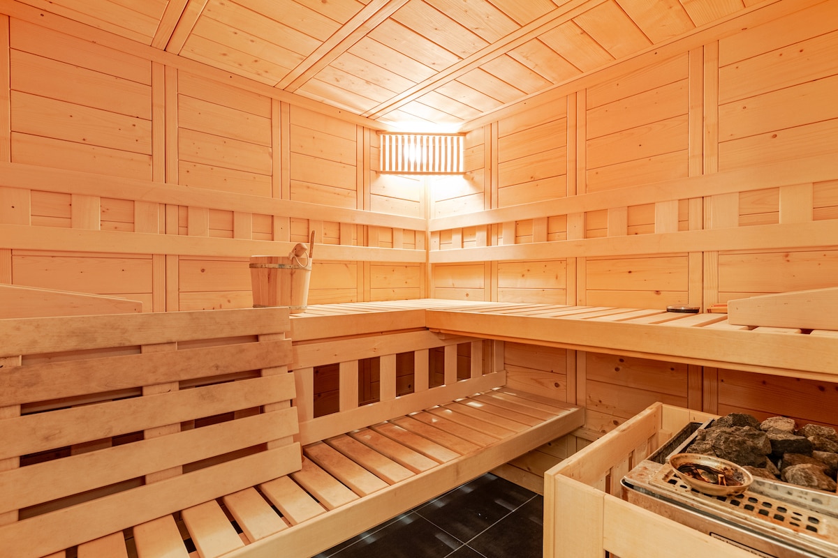 A wooden sauna interior features warm-toned paneling, accentuated by soft lighting. Bench seating is arranged along the walls, with a traditional sauna stove placed prominently. A small wooden bucket is visible, adding to the authentic sauna experience.