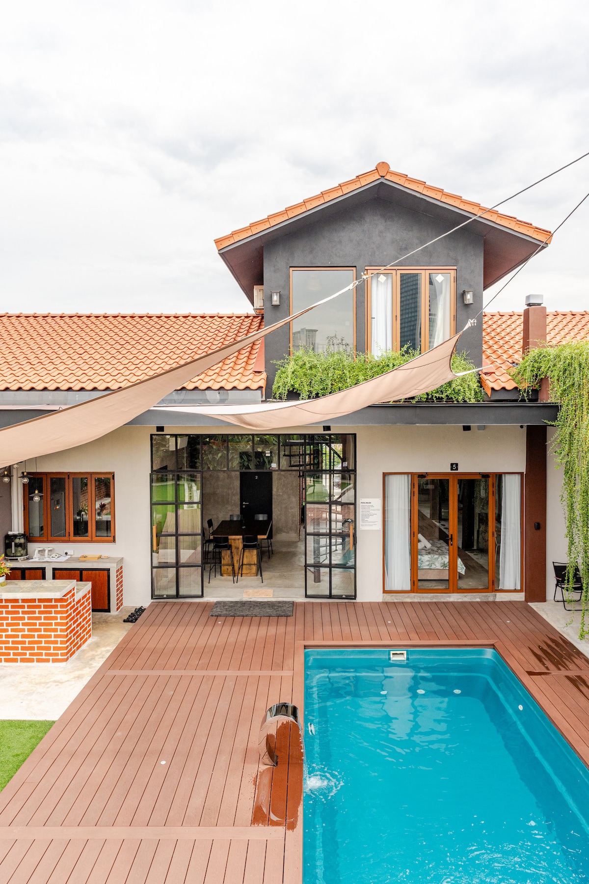 A modern villa is depicted, featuring a wooden deck surrounding a clear blue pool. The house has large glass doors leading to a spacious interior, adorned with subtle earthy tones. A shaded outdoor area is highlighted, complemented by lush greenery and a terracotta roof.