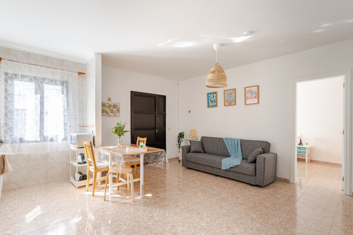 A spacious living area is shown, featuring a grey sofa with a light blue blanket draped over it. A small dining table with four wooden chairs is positioned nearby. Large windows allow natural light to fill the room, and decorative elements adorn the walls.