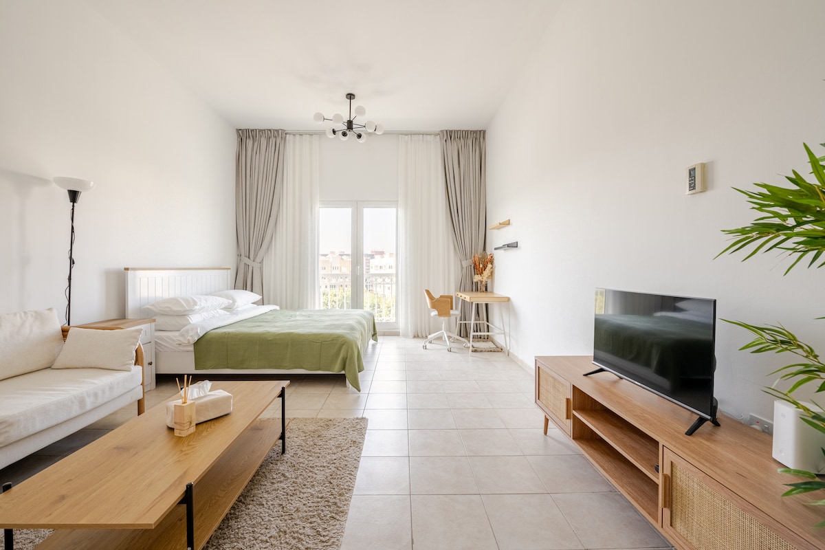 A bright studio space features a spacious bed with light bedding and a green blanket. A small dining area is visible alongside a styled desk and chair. Natural light fills the room through large windows, complemented by light curtains. Modern furnishings and a minimalist decor enhance the open feel.
