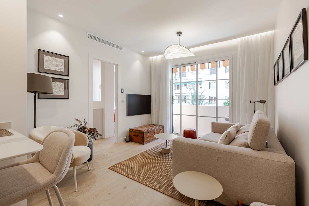 A modern living area features a comfortable sofa and a wooden coffee table, with a light-colored rug underneath. Natural light enters through large windows, accented by an electric awning. A wall-mounted TV and framed artwork enhance the space's aesthetic appeal.