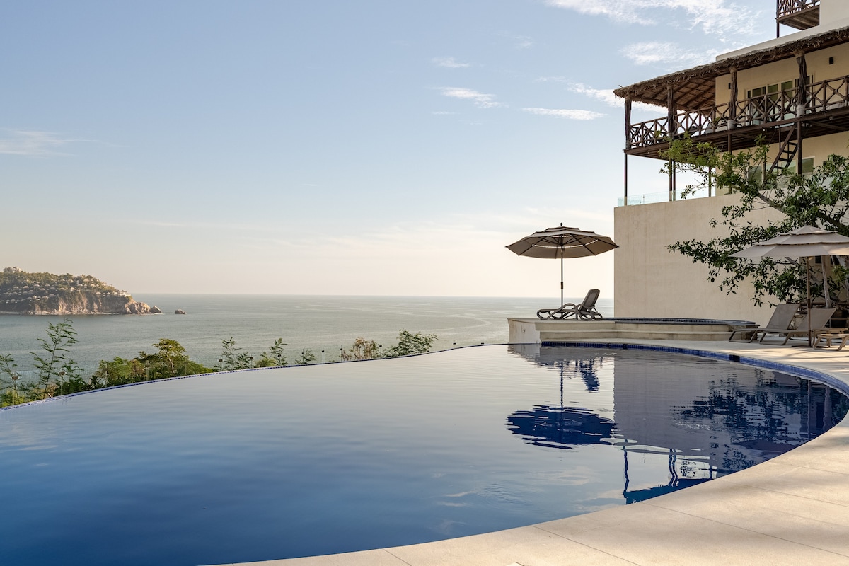 An infinity swimming pool extends towards the horizon, blending seamlessly with the ocean view. A shaded lounge area with an umbrella and a chaise lounge is situated nearby, while the surrounding landscape features gentle slopes and distant hills.