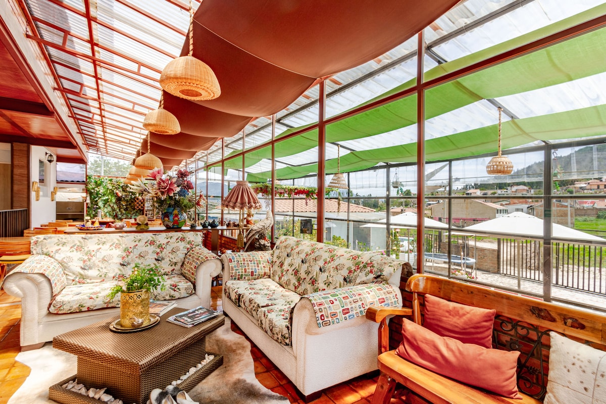 A spacious lounge area is featured, adorned with floral-patterned sofas and wooden seating. Abundant natural light filters through the glass roof, complemented by colorful canopies. Decorative plants and light fixtures enhance the inviting ambiance, while the outdoor scenery is visible through large windows.