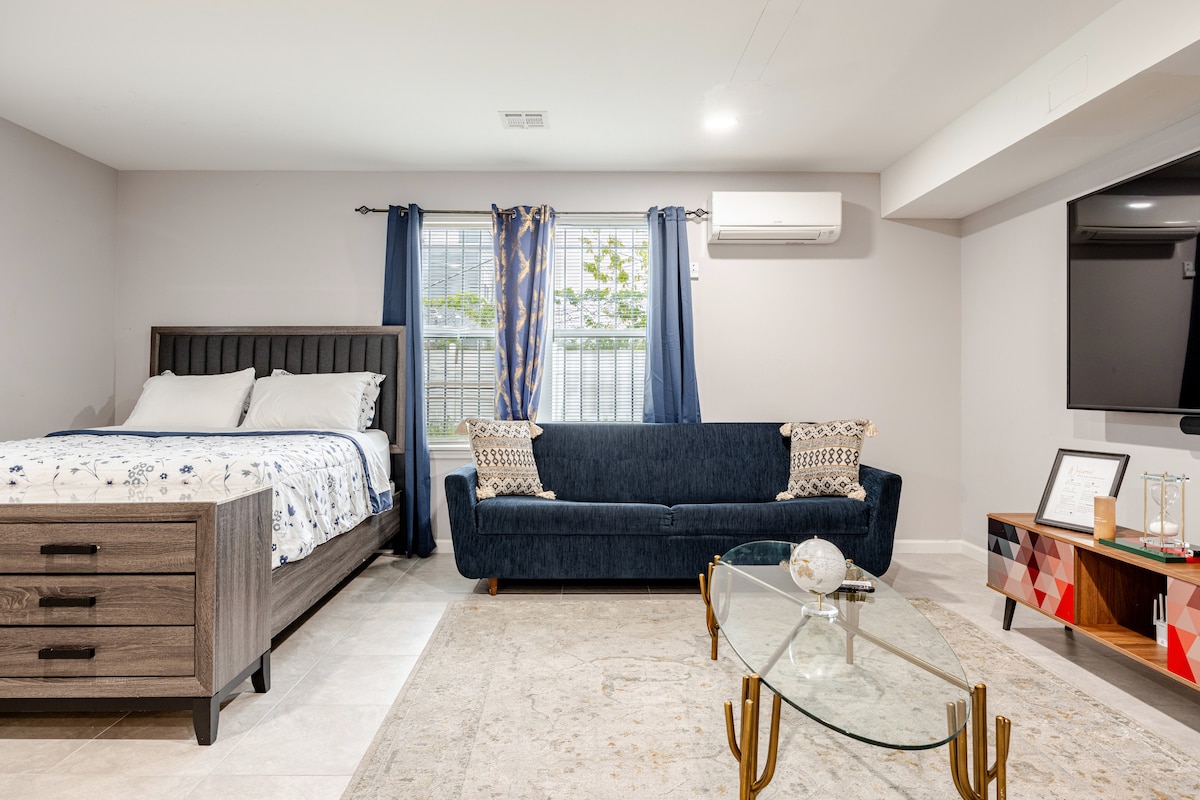 A modern studio features a spacious bed with a decorative headboard and light bedding, situated near a window dressed with blue curtains. A cozy dark blue sofa complements the room, alongside a stylish coffee table and a flat-screen TV mounted on the wall.