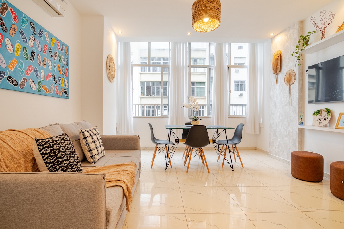 A modern living space features a light grey sofa with a decorative throw. A glass dining table is surrounded by black chairs, while a colorful artwork hangs on the wall. Natural light fills the room through large windows, complemented by neutral-toned flooring.