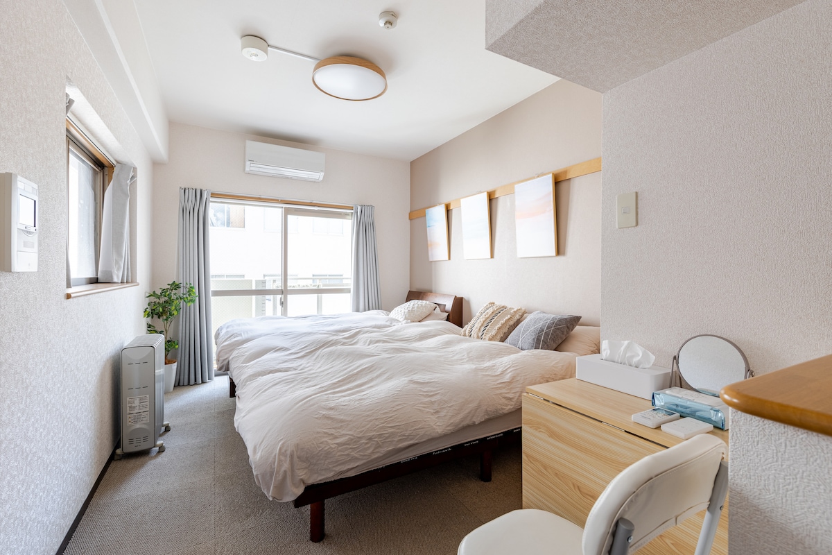 A bright and airy room features two spacious beds dressed in white linens. Soft natural light filters through the sliding glass door, leading to a small balcony. A wooden desk and chair are positioned nearby, complemented by a modern wall-mounted light fixture.