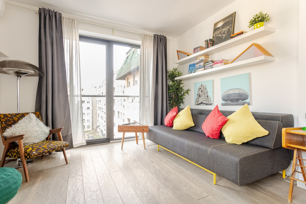 A bright living area features a gray sofa adorned with colorful cushions. A stylish wooden coffee table sits in front, and a nearby chair adds to the cozy ambiance. Light enters through a large window, complemented by soft curtains, while books and decor adorn the wall-mounted shelves.