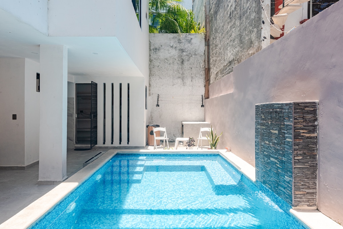 A private pool area is showcased, featuring clear blue water surrounded by a clean, modern layout. White sun loungers are positioned nearby, and decorative plant life adds a touch of greenery. The surrounding walls are finished in light tones, enhancing the bright atmosphere of the space.