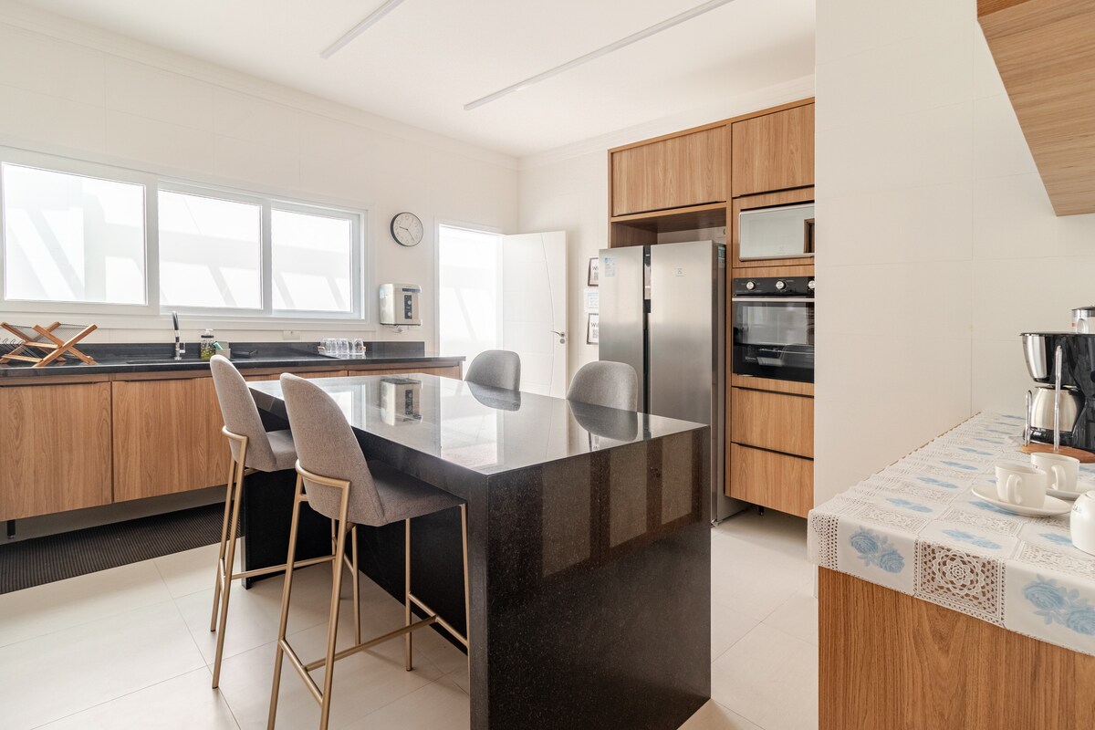 A modern kitchen features sleek cabinetry and a large kitchen island with seating for three. Stainless steel appliances, including an oven and refrigerator, are visible. Natural light floods the space through multiple windows, enhancing the minimalist design.