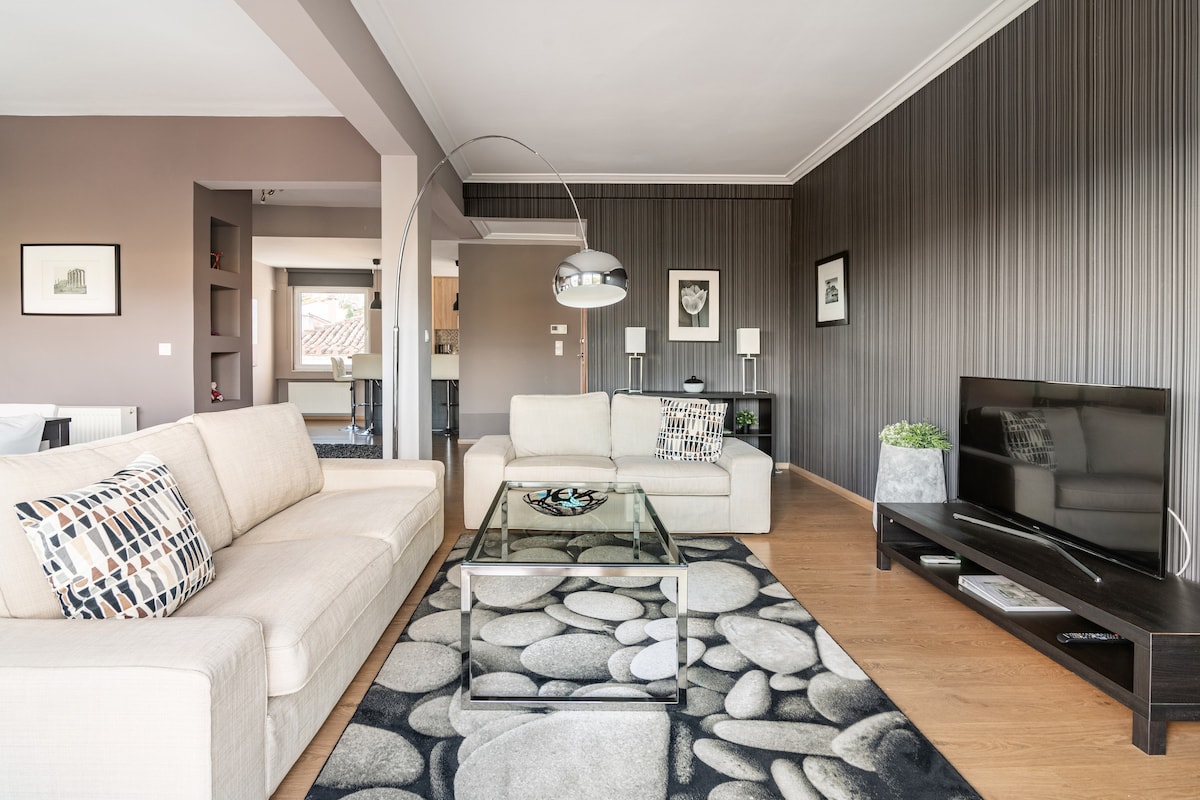 The spacious living room features two comfortable couches and a modern coffee table placed on a decorative rug. A large TV is positioned on a wooden console, while subtle wall decor adds to the room's style. Natural light fills the space through nearby windows.