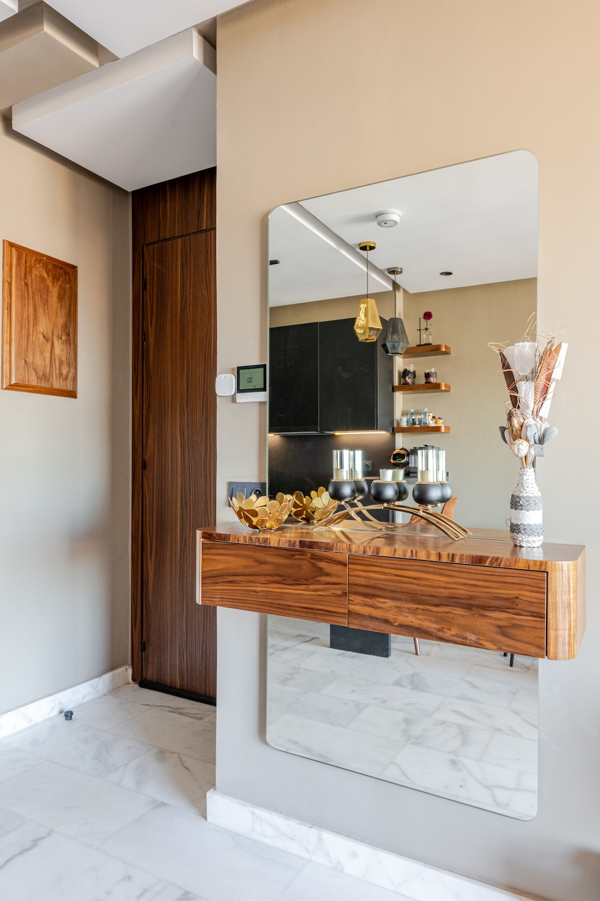 A modern entryway features a sleek wooden console with two drawers and a large mirror above. A minimalist decor is complemented by a vase with dried flowers. The backdrop showcases an open kitchen with contemporary shelving, illuminated by pendant lighting.