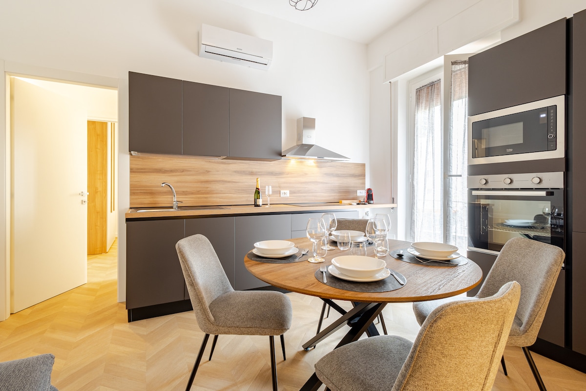 A modern kitchen area features sleek cabinetry accompanied by a wooden countertop and stainless steel appliances. A round dining table is set for four, and large windows allow natural light to brighten the space. The open layout connects seamlessly to a hallway leading to other rooms.