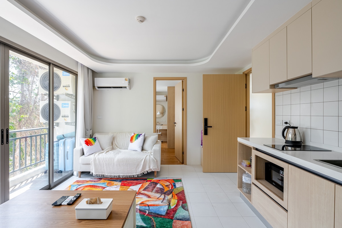 A light-filled living area features a modern couch adorned with colorful cushions. A vibrant area rug adds warmth to the space. Open shelving holds a kettle and microwave, while large glass doors provide access to a balcony, enhancing indoor-outdoor connectivity.