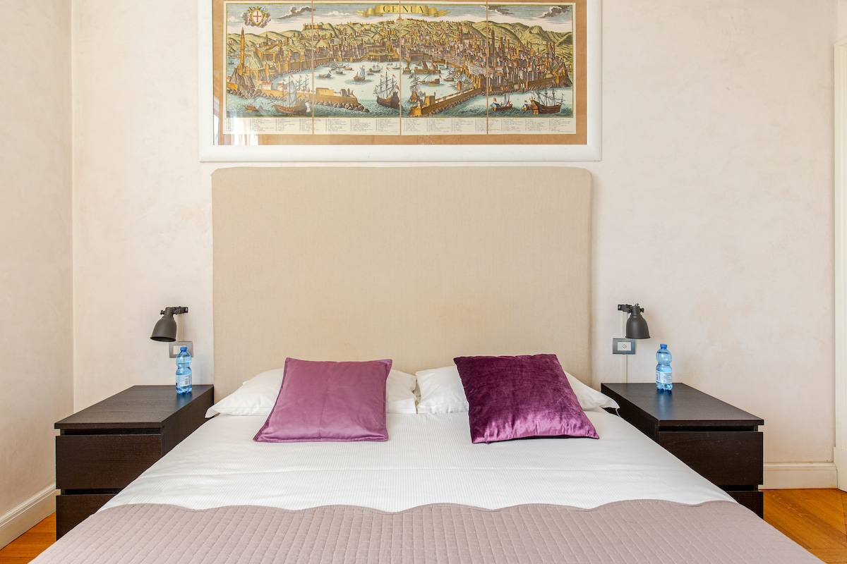 A spacious bed is positioned in the center, adorned with two purple accent pillows. Dark wooden nightstands are placed on either side, featuring modern lamps. Above the bed, a vintage map artwork adds a historical touch to the light, neutral-toned wall.