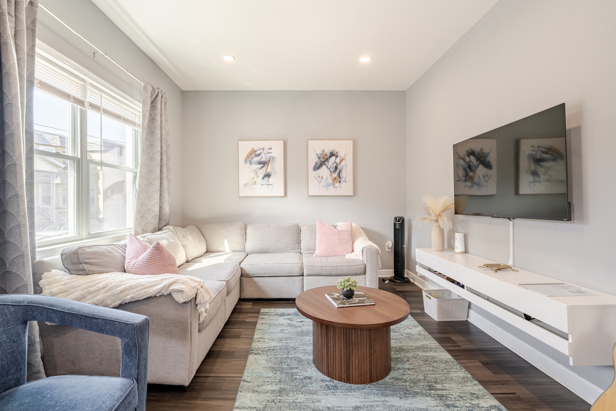 The living room features a spacious sectional sofa adorned with soft pillows, providing ample seating. A round wooden coffee table sits at the center, surrounded by light carpeting. Two framed artworks decorate the walls, and a mounted smart TV enhances the space, supplemented by natural light from the window.