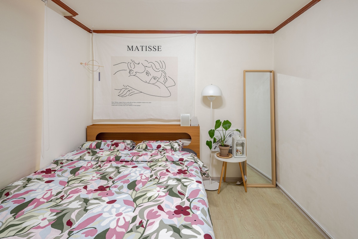 A bright sleeping area features a spacious bed adorned with a floral-patterned comforter. A soft light stands beside the bed, while a mirror and a small side table with plants are positioned nearby. A piece of wall art adds a touch of creativity to the room.