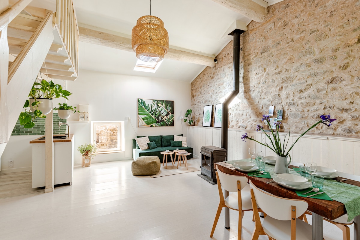 A spacious living area features exposed stone walls and light wooden beams. A green sofa and neutral decor offer comfort, while a large skylight allows natural light to fill the room. A dining table with four chairs is set, accompanied by decorative plants and artwork.