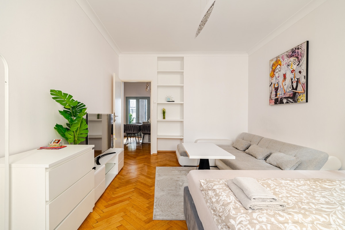 A bright living area is presented with a spacious gray sofa and a minimalist coffee table. A television stand is featured against one wall, while a light wood floor adds warmth. A decorative plant provides a touch of greenery next to the modular shelving.