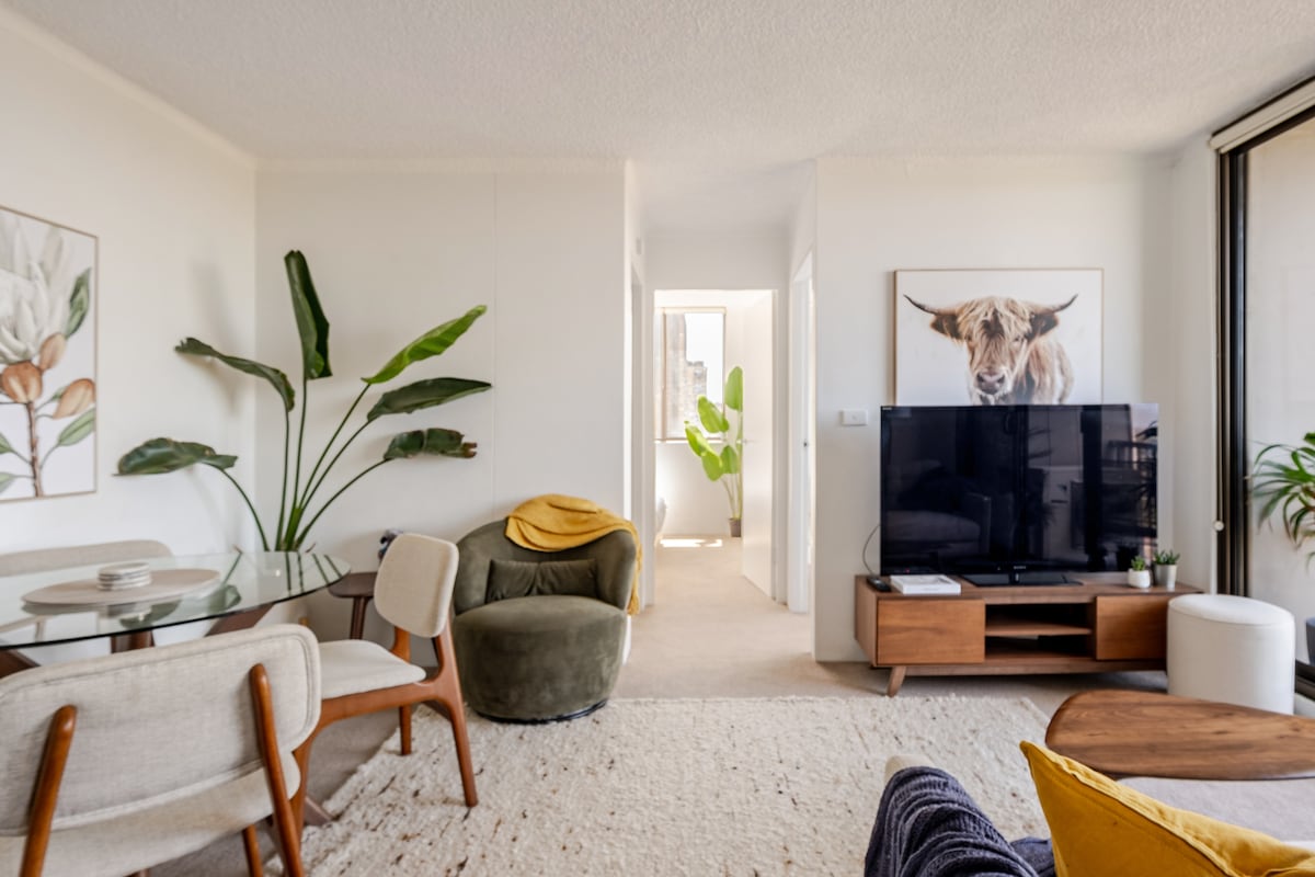 A comfortable living area is featured, with a mounted television on a wooden unit. A round coffee table and a dining table with chairs are arranged. Lush green plants add a touch of nature, while warm tones are introduced by a cozy throw on a chair.