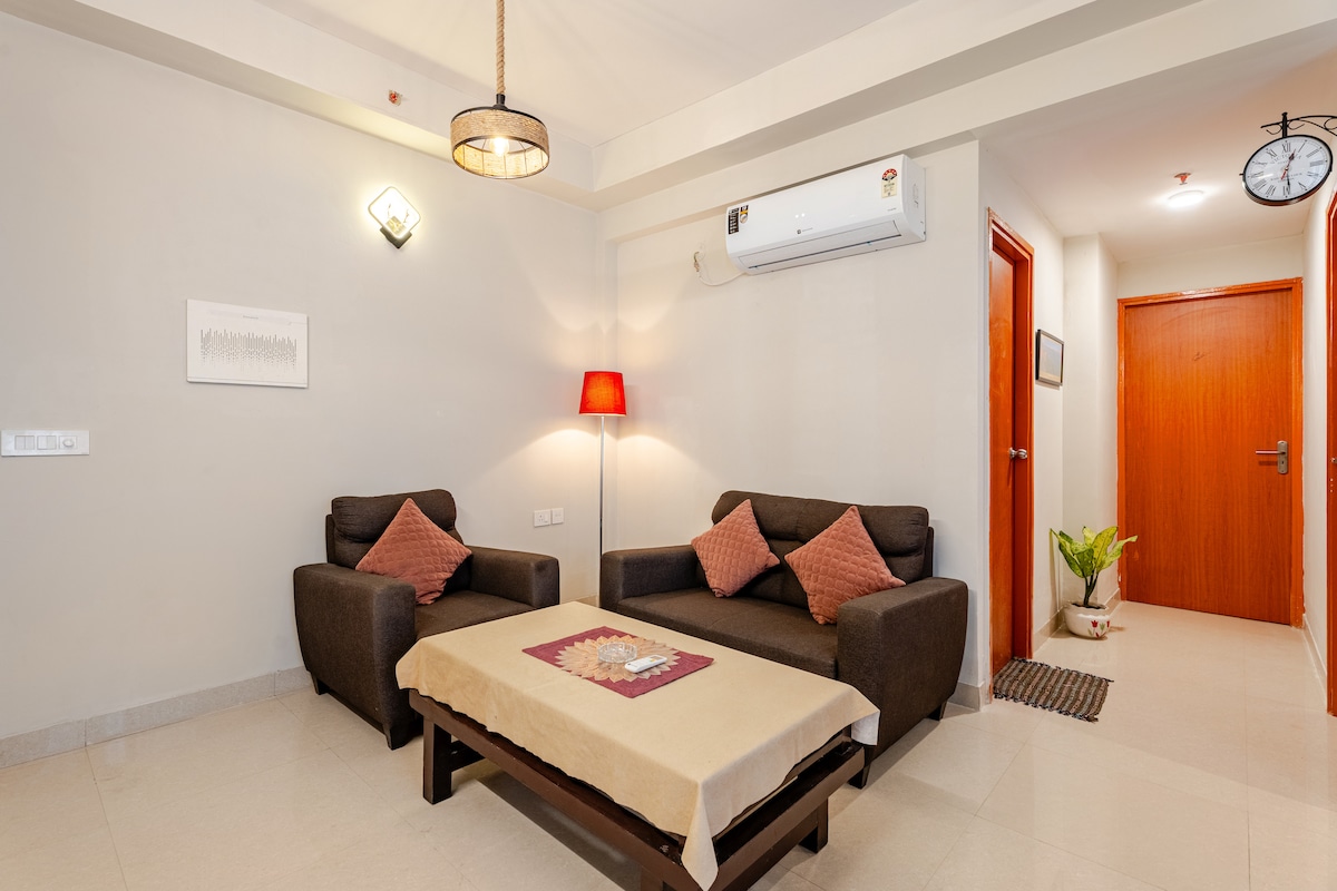 A living area is featured with two dark armchairs and a coffee table positioned in front. A table lamp with a warm glow illuminates the space. A wooden door and a clock are visible in the background, along with a small plant adding a touch of greenery.