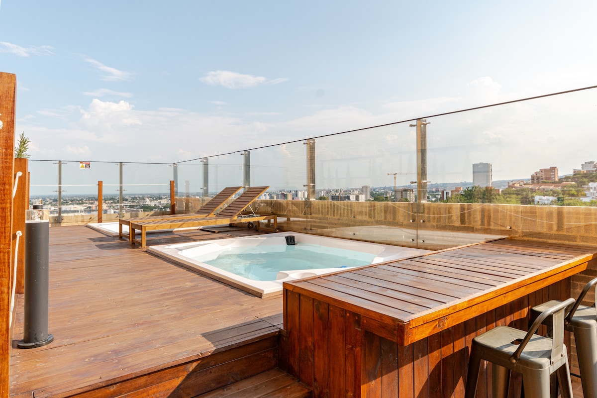 An expansive rooftop terrace features a wooden deck surrounding a circular jacuzzi. Lounge chairs are positioned for relaxation, offering panoramic views of the city skyline and distant landscape. A wooden bar area with high stools complements the outdoor space, providing a perfect spot for refreshments.
