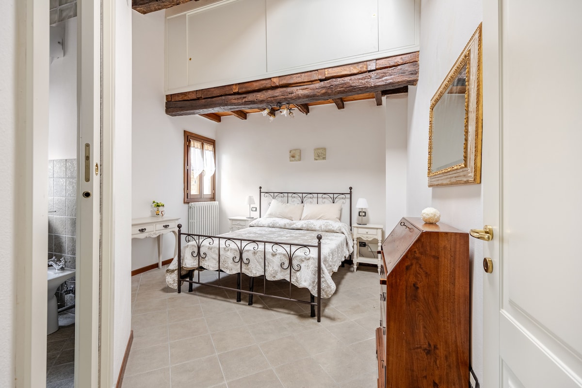 A bright bedroom is presented with wooden beams overhead and soft tones. A spacious bed is centered, dressed with light bedding. A wooden chest and a small table are featured, with natural light filtering through the window, creating a serene environment. A mirror hangs on the wall.