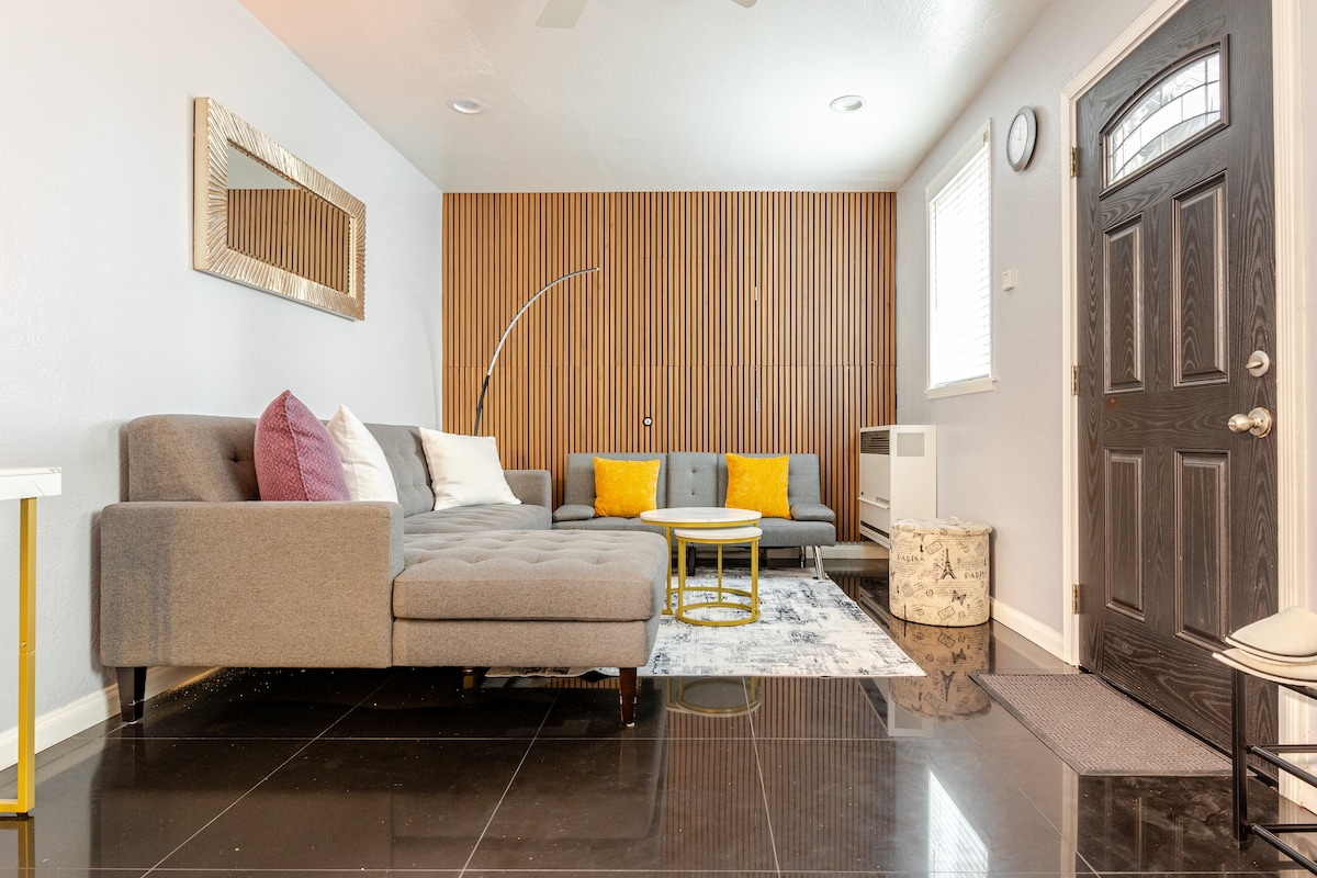 A modern living room features a gray sectional sofa with colorful pillows arranged around a circular coffee table. A decorative rug lies on shiny black tiled flooring, and natural light enters through a window with blinds. A stylish accent wall and a welcoming front door add character.