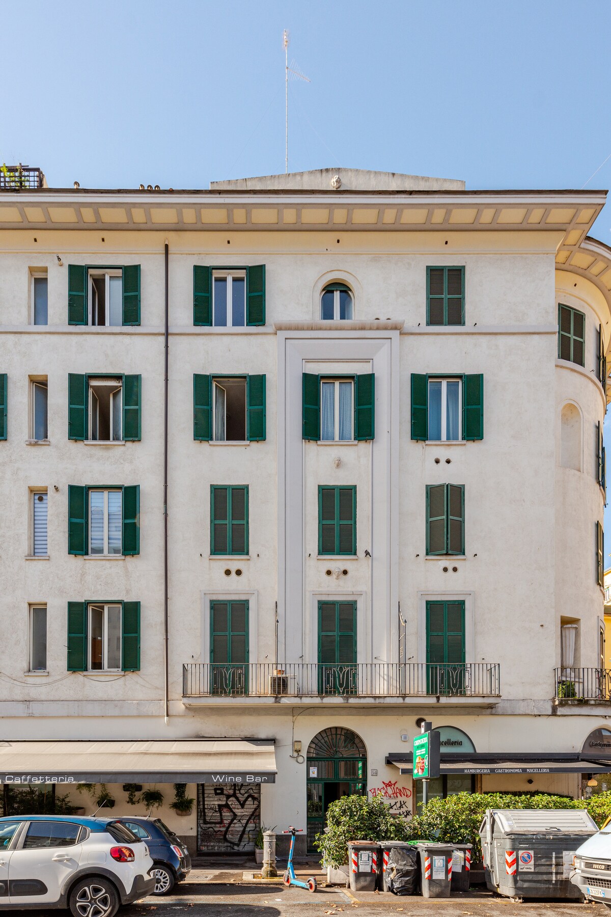 The exterior of a three-story building is presented, featuring white walls and green shutters adorning the windows. A balcony extends from the second floor, and a small entrance is visible on the ground level. Partial views of nearby shops and street activity are included.