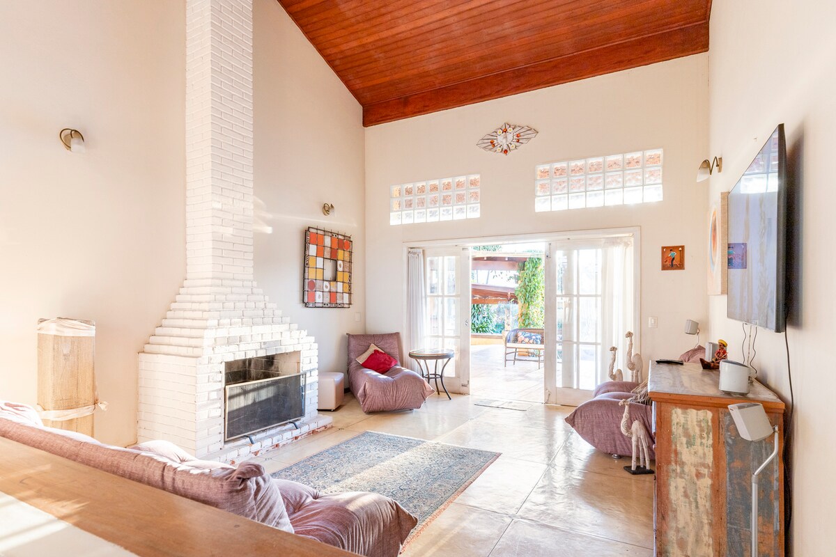 A spacious living area features high ceilings with wooden beams and a white brick fireplace as a focal point. Large glass doors allow natural light to enter, creating a bright atmosphere. Comfortable seating options and a decorative rug add a cozy touch to the space.