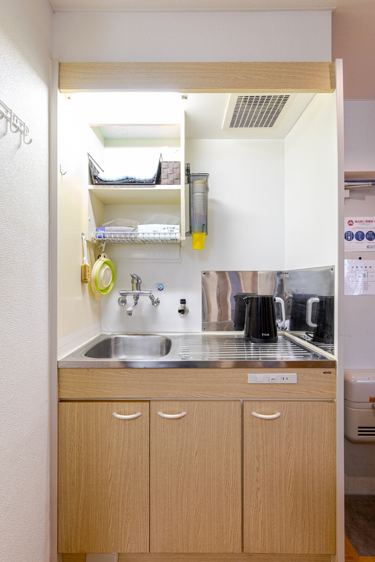 A compact kitchenette is presented with wooden cabinets and a stainless-steel sink. Above, shelves hold kitchen essentials, while a kettle and other appliances are visible on the countertop. The space is designed for convenience, with ample storage for culinary needs.