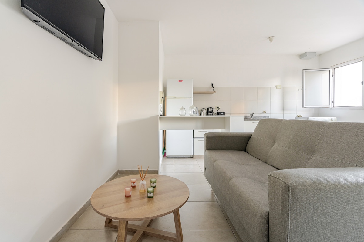 A light-filled living area with a spacious gray sofa is positioned in front of a mounted TV. A round wooden coffee table rests on the tiled floor, complemented by soft lighting from a nearby window. The kitchen area is visible in the background, featuring modern appliances.