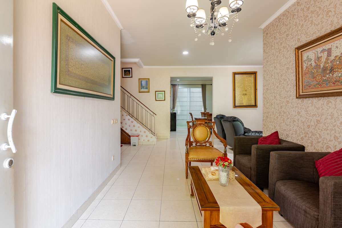 An open living area showcases a light tiled floor, complemented by framed artwork on the walls. A wooden coffee table sits at the center, surrounded by plush seating. A staircase is visible in the background, leading to the upper level of the house.