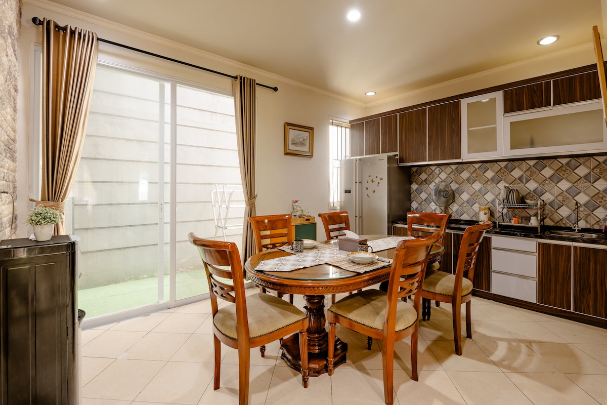 A dining area is showcased with a round wooden table surrounded by six chairs. Natural light enters through the sliding glass door, enhancing the space's openness. A compact kitchen with wooden cabinetry and modern appliances is visible in the background, providing a functional and inviting atmosphere.