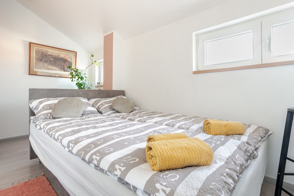 A cozy sleeping area is highlighted by a spacious bed with patterned bedding and decorative pillows. Two rolled towels are neatly placed at the foot of the bed. A window allows natural light to fill the room, complemented by a green plant on the nearby surface.