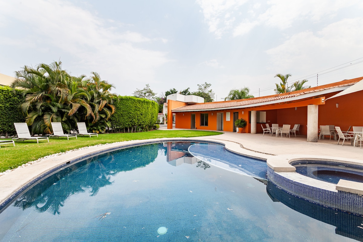 A private swimming pool with a circular design is surrounded by a lush green lawn. Sun loungers are positioned near the pool, and a jacuzzi is visible next to the water. The vibrant orange walls of the house complement the tranquil outdoor setting.