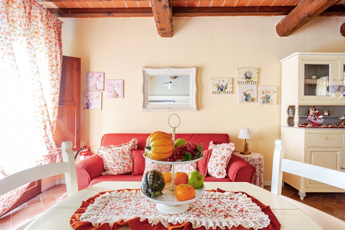 A bright living area features a red sofa adorned with floral cushions, set against warm yellow walls. A round table is presented with a fruit centerpiece, surrounded by white chairs. Decorative pieces and artwork are displayed on the walls, with natural light filtering through floral curtains.