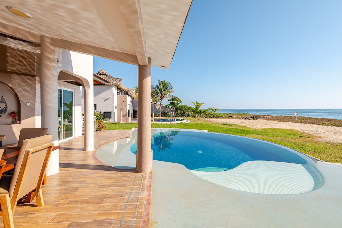A serene outdoor space features a curved swimming pool with a clear blue surface, surrounded by well-maintained grass. Beach access is visible in the distance, and palm trees provide a natural backdrop, creating a tranquil environment that invites relaxation.
