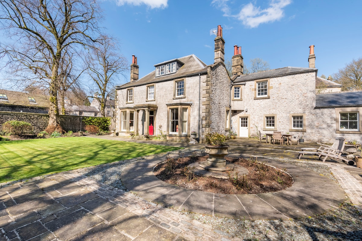 The exterior of the Old Vicarage is showcased, featuring a charming stone façade and well-maintained garden. A circular stone pathway is seen surrounding a central garden bed. Outdoor seating is available, complemented by large windows, allowing natural light to enhance the atmosphere.