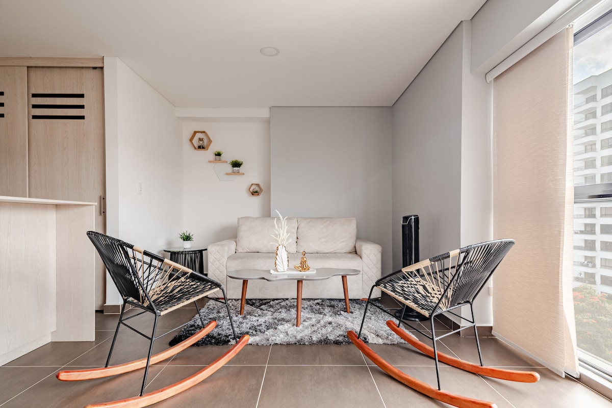 A modern living area features two rocking chairs with black and natural wood finishes. A light-colored sofa is positioned across from a simple coffee table. Plants are displayed on a wall shelf, complementing the neutral tones of the walls and flooring.